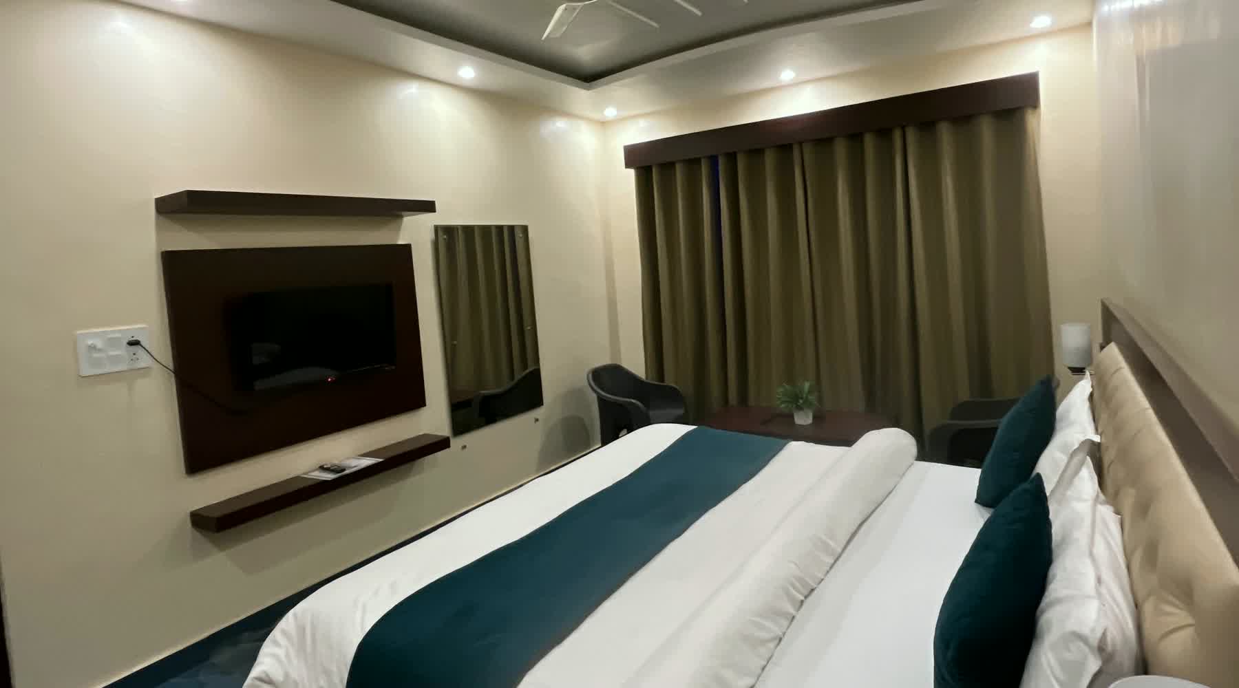 A guest room with a teal runner on the bed, a wall-mounted television and long curtains.